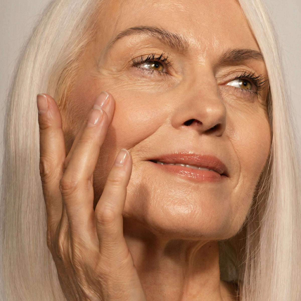 Close-up of a smiling older woman with long white hair touching her cheek gently, showing smooth skin and natural makeup, against a neutral background.