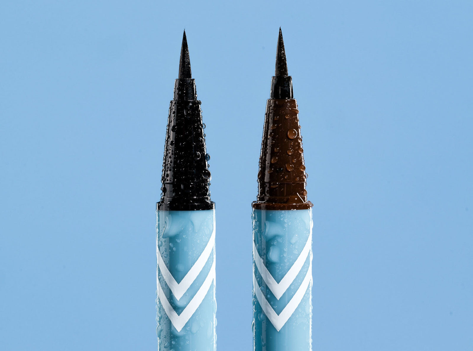 Two blue liquid eyeliner pens with fine tips and white chevron patterns stand upright against a light blue background, both covered in water droplets.