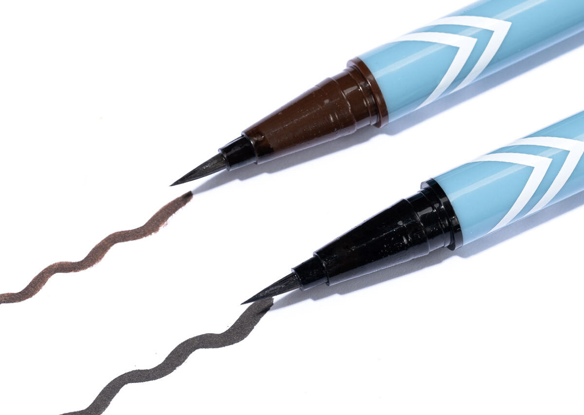 Two light blue felt-tip eyeliner pens with pointed tips draw wavy lines, one brown and one black, on a white surface.