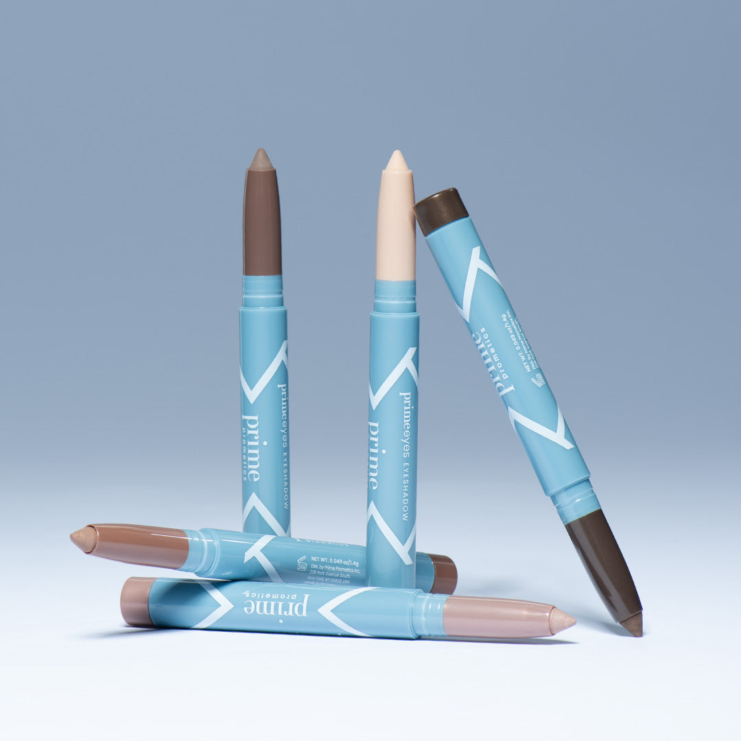 Four dual-ended makeup sticks in light blue packaging are standing and lying on a light surface with a gradient blue background. Each stick features different shades of brown and beige on the ends.