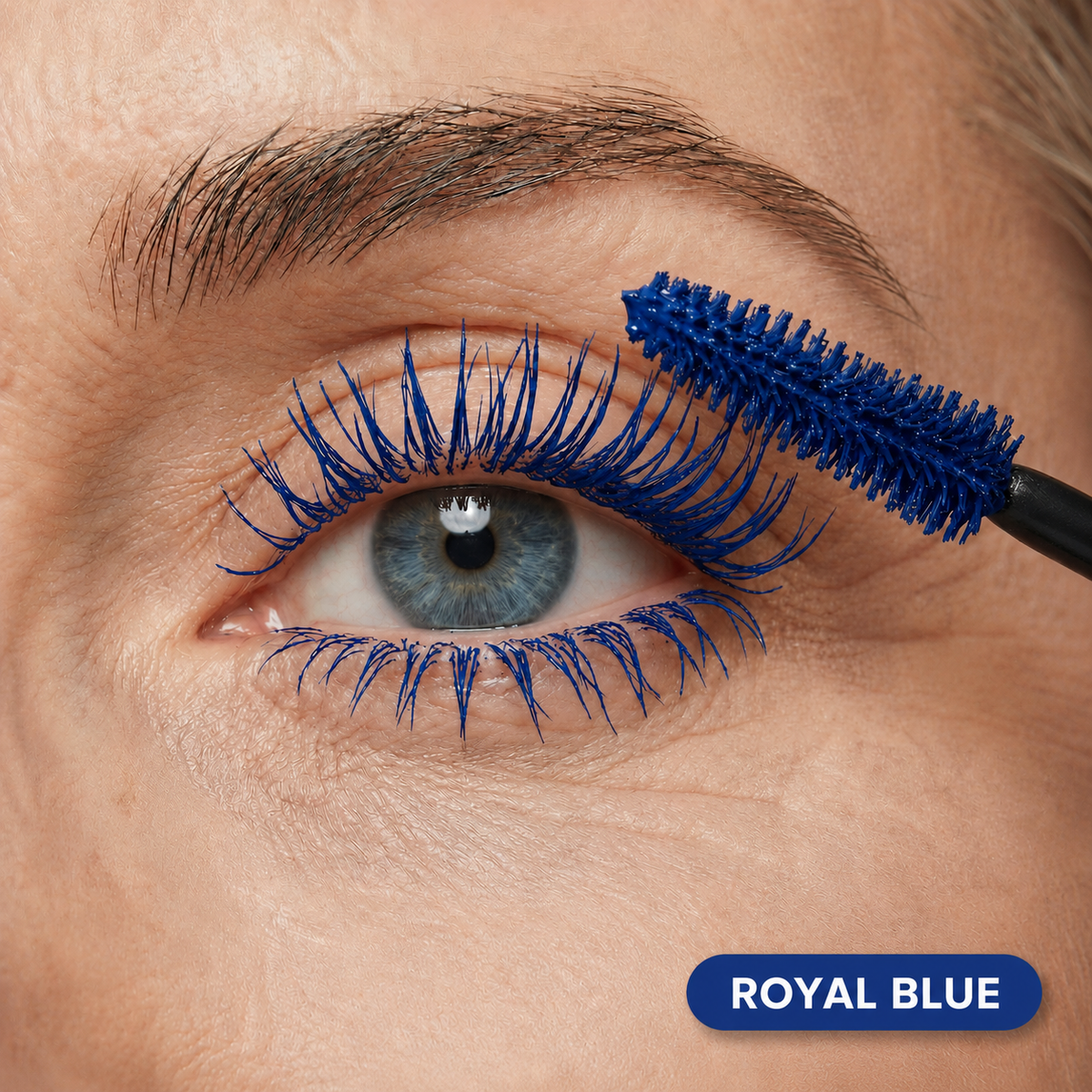 Eine Nahaufnahme eines Auges, bei der die PrimePrometics™ PrimeLash Mascara in Königsblau auf die oberen Wimpern aufgetragen wird. Der Mascara-Applikator ist zu sehen, und auf einem Etikett in der unteren rechten Ecke steht „ROYAL BLUE“. Diese speziell für reife Frauen entwickelte Pro-Age-Mascara fördert das Wimpernwachstum und sorgt gleichzeitig für eine kräftige, leuchtende Farbe.