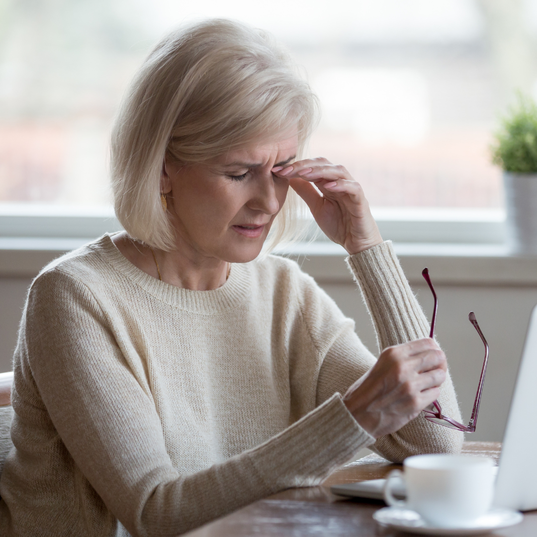An older woman with short blonde hair sits at a table holding her glasses in one hand and pressing her forehead with the other, appearing to have a headache or be stressed. A laptop and coffee cup are on the table.
