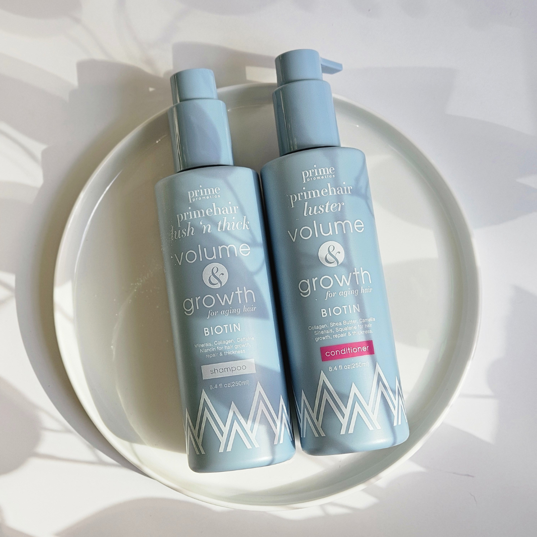 Two light blue bottles of Prime Primehair shampoo and conditioner labeled “Volume & Growth” sit on a white plate in bright, natural light with soft shadows and abstract shapes in the background.