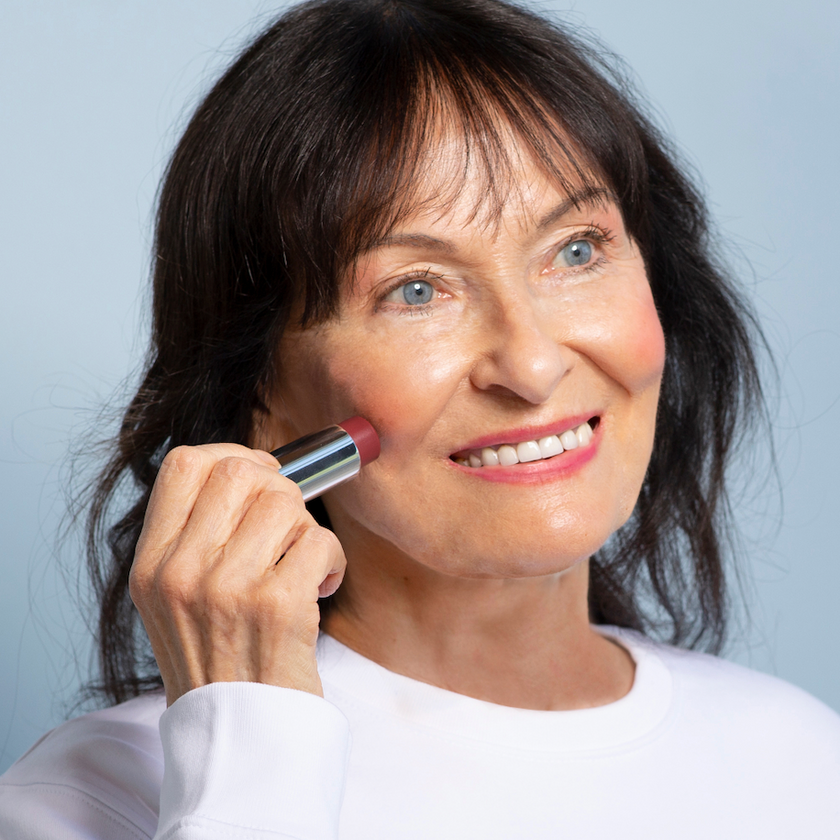 Smiling older woman with blue eyes and dark hair applies pink blush to her cheek using a makeup stick, wearing a white top against a light blue background.