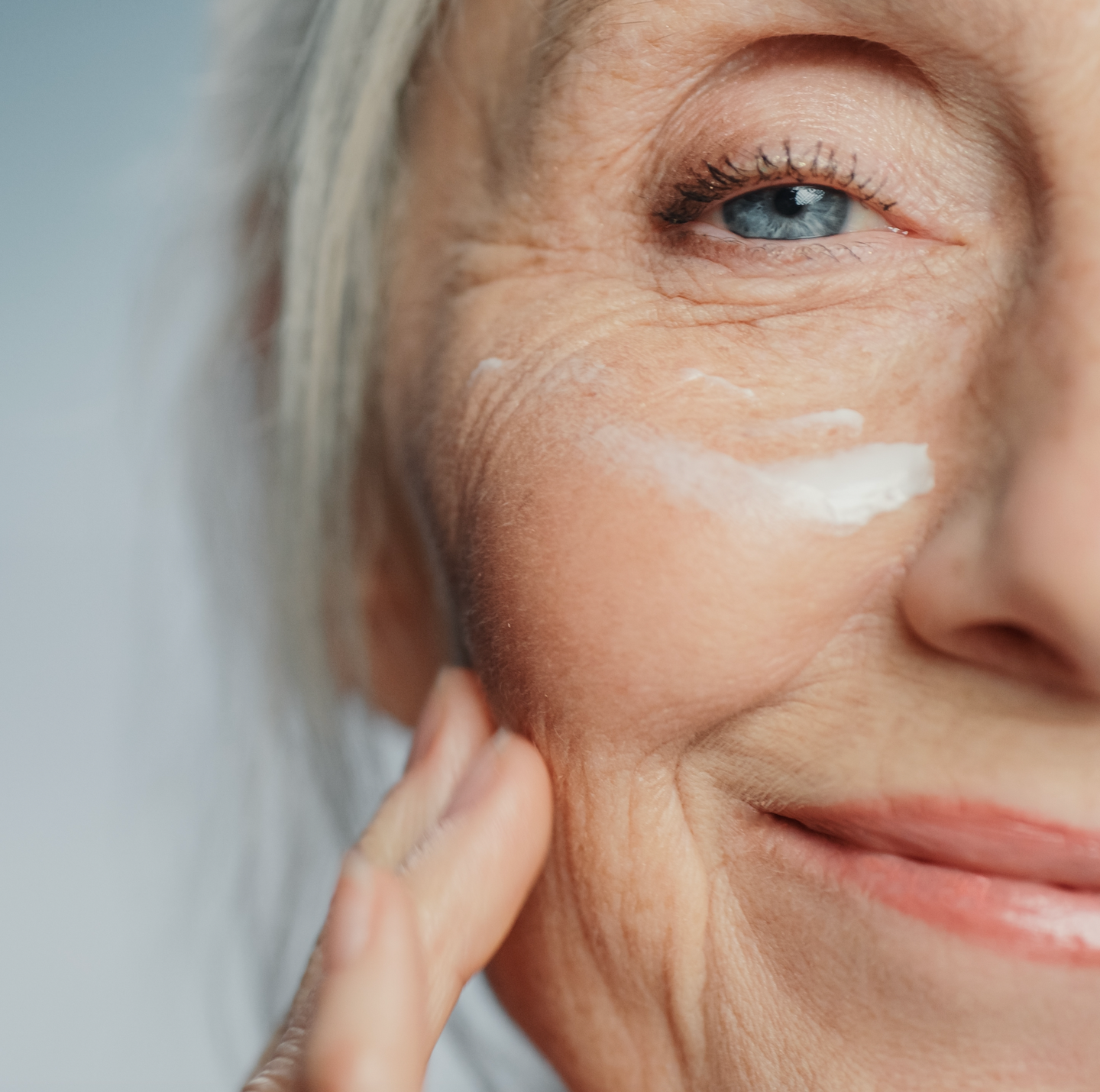 Close-up of an older womans face, smiling, with a visible blue eye and gray hair. She is applying a streak of white cream to her cheek with her finger, highlighting her smooth skin and fine lines.