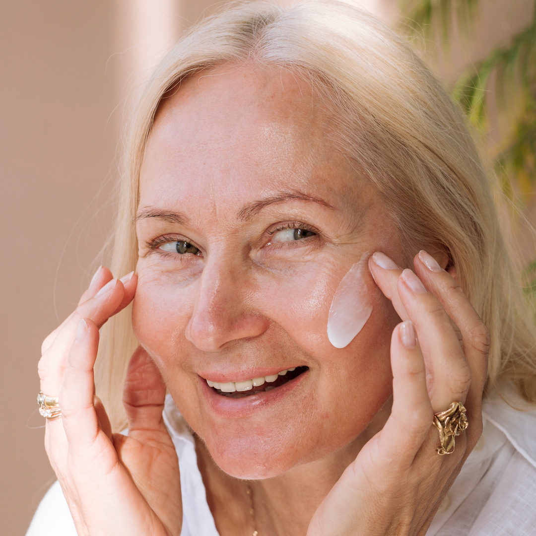 Smiling older woman with light hair applies a cream to her cheek with both hands. She wears a white top and gold rings, and stands in soft natural light with greenery in the background.
