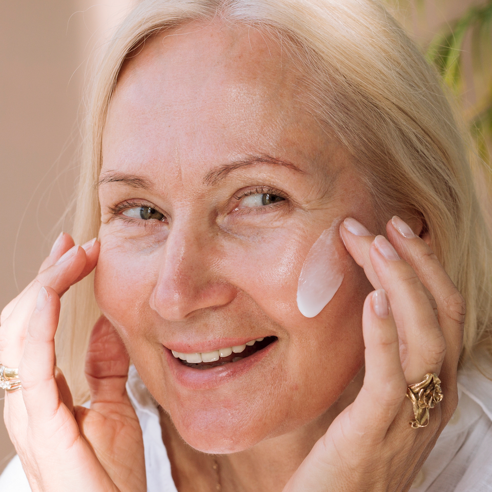 Smiling older woman with light hair applies a dab of skincare cream to her cheek with her fingertips. She wears a white top and gold rings, and appears relaxed and happy.