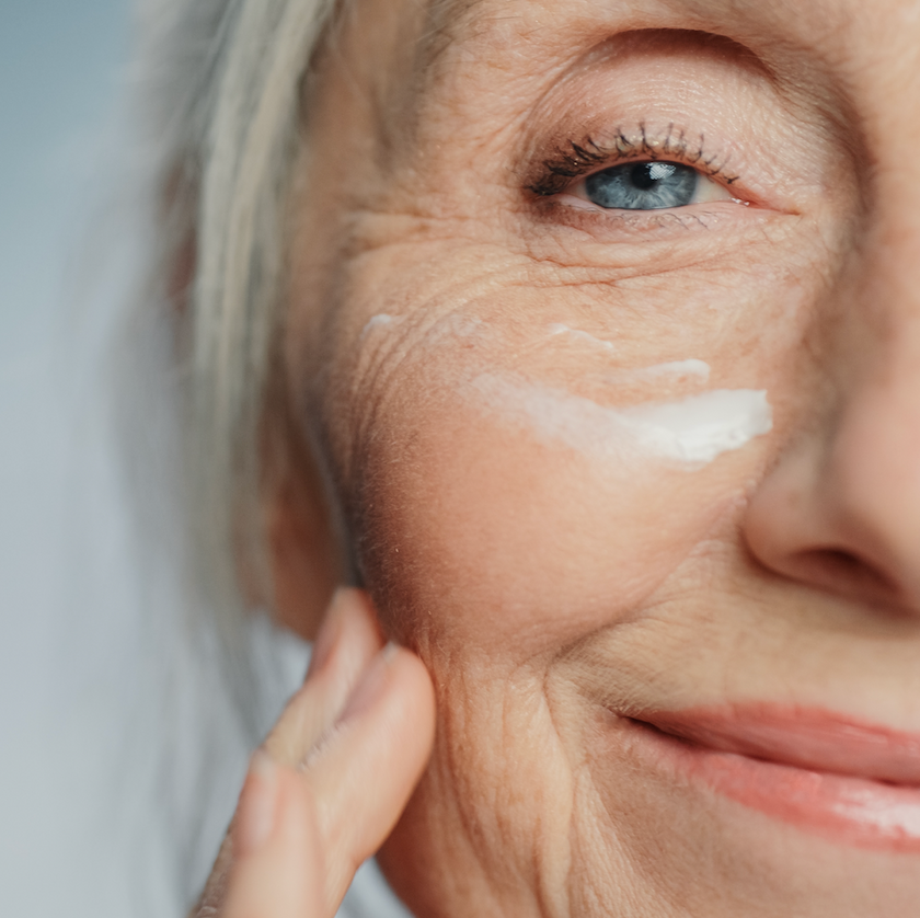 Close-up of an older woman smiling and applying cream to her cheek, with visible wrinkles and blue eyes, suggesting skincare or self-care.