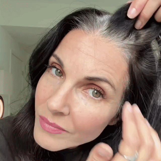 A woman with dark hair and green eyes shows her gray roots by lifting a section of her hair, looking directly at the camera in a well-lit room.