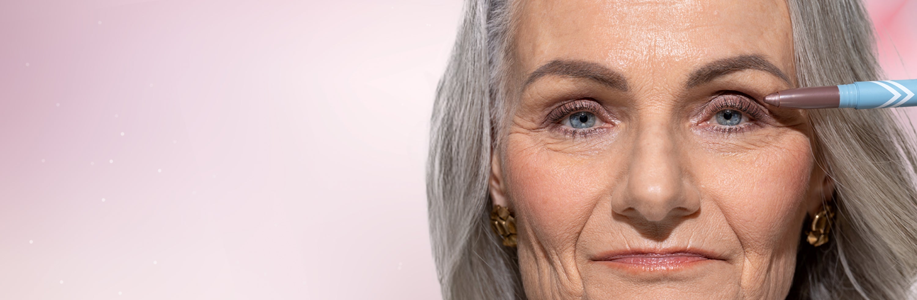 Close-up of an older woman with gray hair and blue eyes, looking at the camera. She is applying makeup with a pencil near her right eye against a soft pink background.