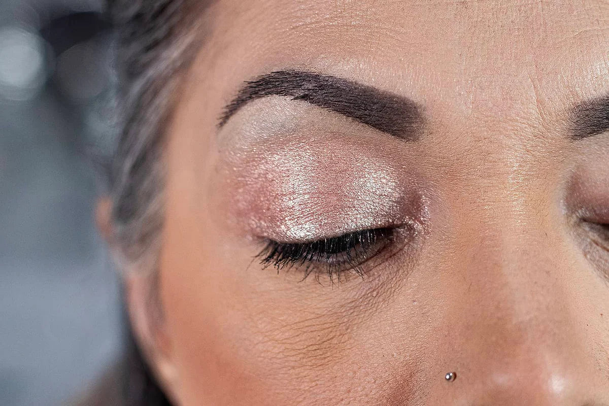 A close-up of a persons face showing one closed eye with shimmery pink eyeshadow, neatly shaped eyebrows, black eyelashes, and a small nose stud. The skin has a smooth, natural complexion.