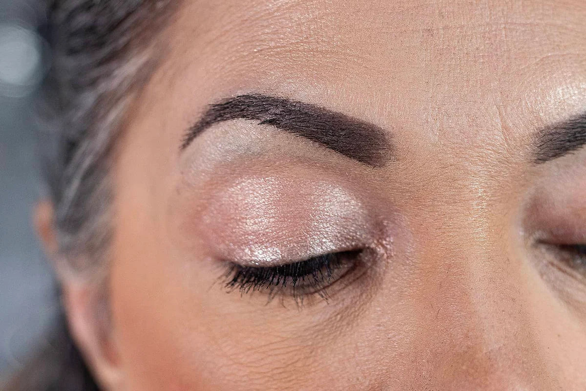 Close-up of a persons closed eye showing shimmery, light pink eyeshadow on the eyelid, well-groomed dark eyebrow, and visible skin texture.