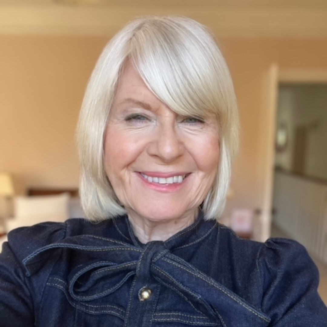 Smiling older woman with straight, shoulder-length blonde hair, wearing a dark denim jacket with a bow at the collar, standing indoors in a softly lit room with neutral walls and some furniture in the background.