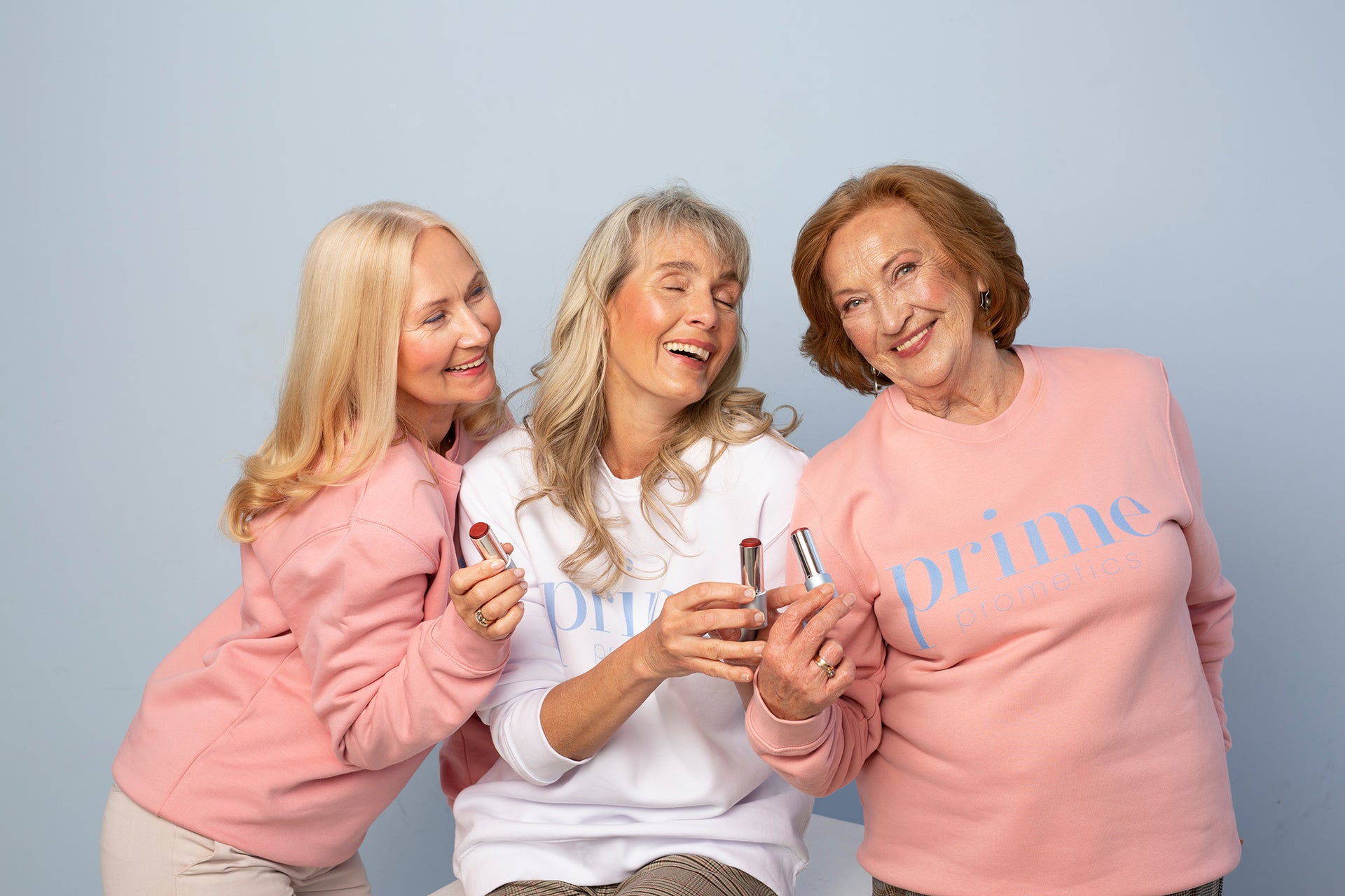 Three smiling older women wearing pastel Prime Cosmetics sweaters hold and display lipsticks against a light blue background, exuding joy and confidence.