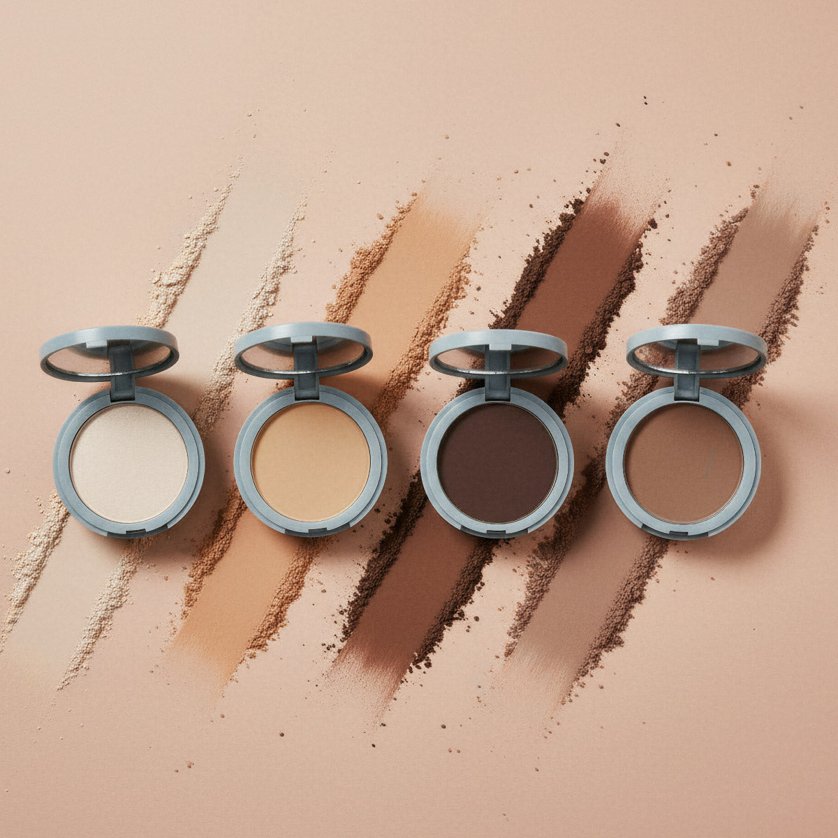 Four open makeup compacts with powders in light to dark shades, each with a matching color swatch brushed diagonally behind them on a beige background.