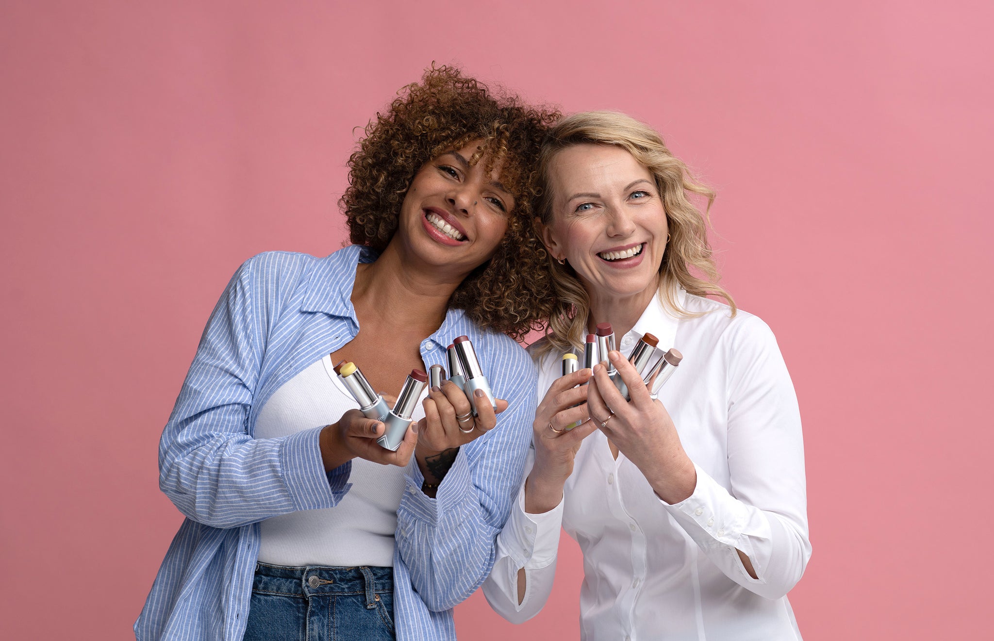Two smiling women stand against a pink background, each holding several lipstick tubes in their hands. They appear happy and are dressed casually in light-colored shirts.