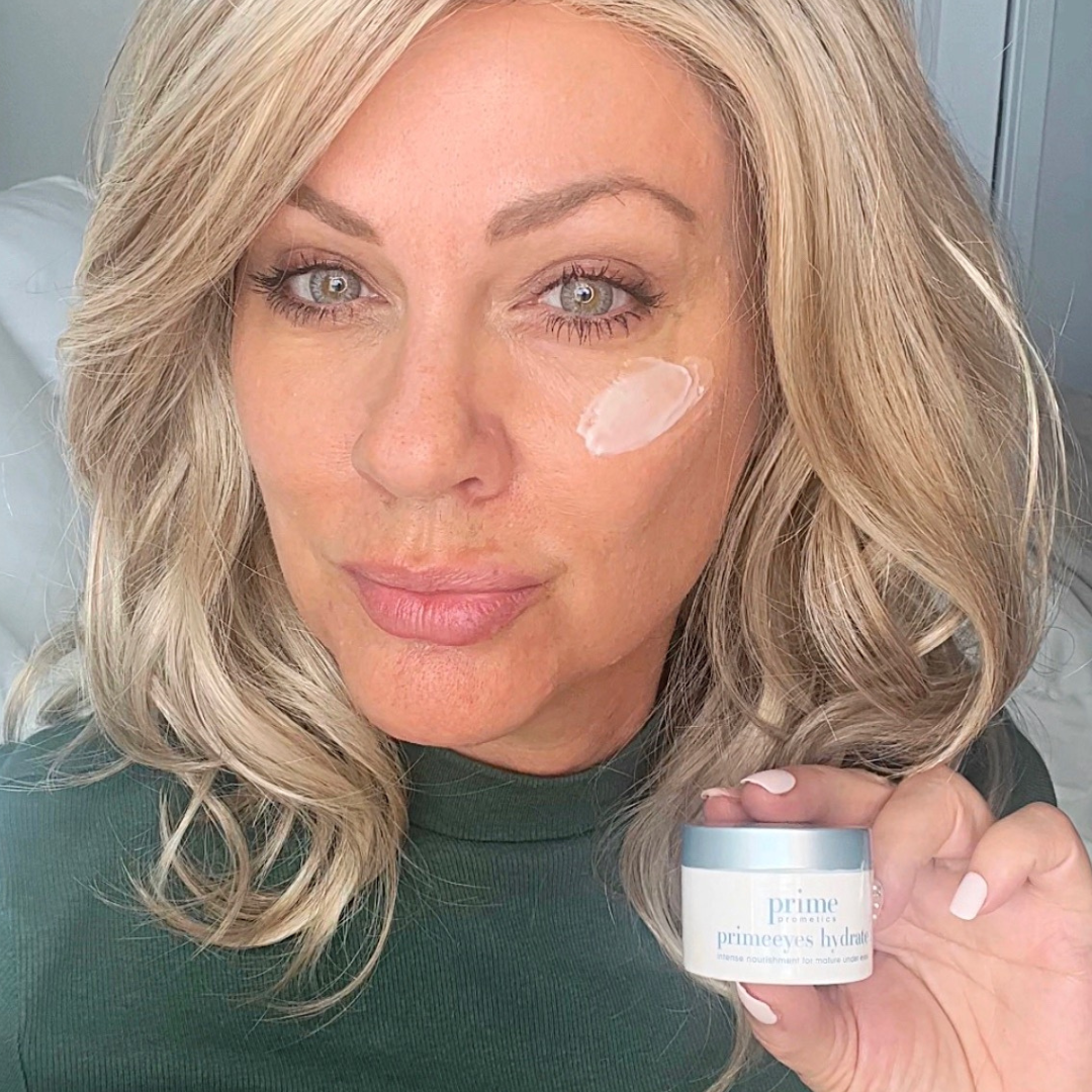 A woman with blonde hair applies a dab of face cream under her right eye and holds a small jar labeled “prime eyes hydra.” She is wearing a green top and looks into the camera.