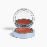 The PrimePrometics™ Undereye Brightening Corrector comes in a circular silver case with a mirror and embossed initials, revealing a warm terracotta shade perfect for targeting dark circles.