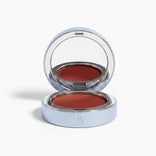 The PrimePrometics™ Undereye Brightening Corrector—a silver compact with mirror—holds a deep reddish-brown balm, perfect for blush, lip color, or as an undereye brightener. Displayed on a white background.
