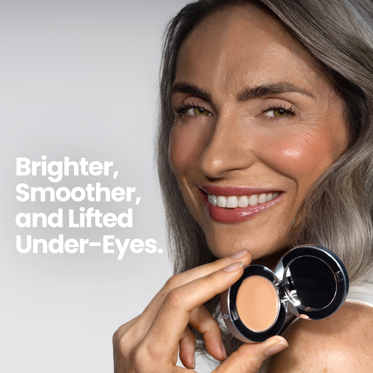 Smiling woman with long gray hair holds an open compact of PrimePrometics™ Undereye Brightening Corrector, revealing radiant skin. Text on image: Brighter, Smoother, Lifted Under-Eyes—ideal for reducing dark circles.
