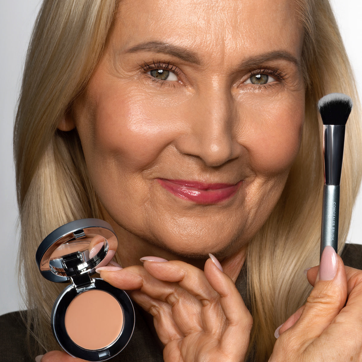 Smiling older woman with long blonde hair holds a makeup brush in one hand and an open compact of cream foundation in the other, demonstrating cosmetic application.