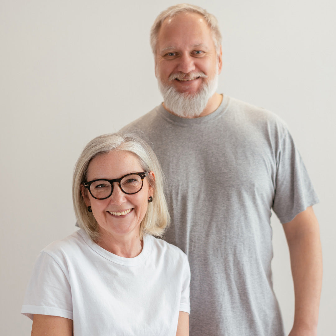 Two older adults, a smiling woman with gray hair and glasses wearing a white t-shirt, and a smiling man with a gray beard wearing a gray t-shirt, stand together against a plain light background.