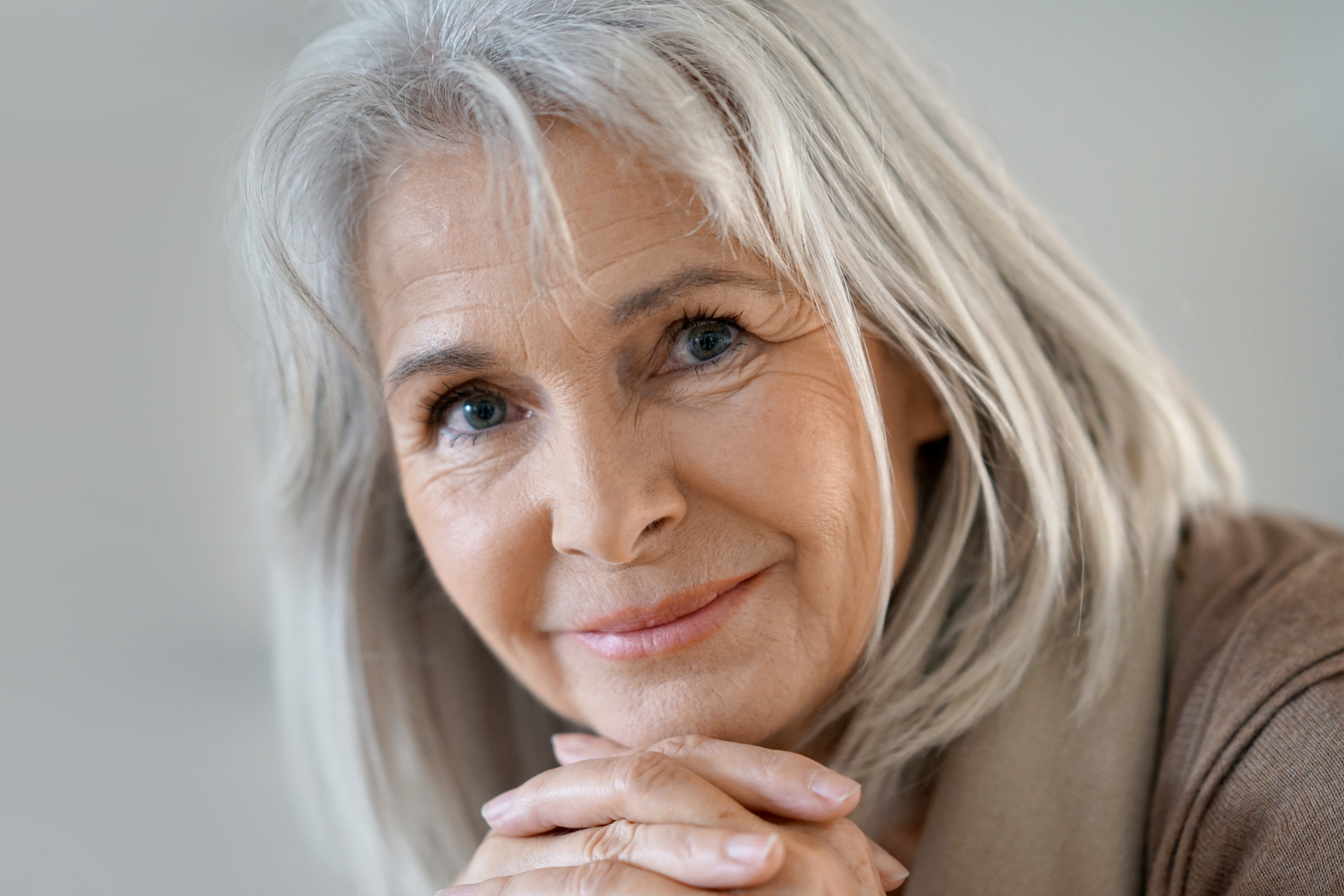 A smiling older woman with gray hair and blue eyes looks at the camera, resting her chin on her hands. She is wearing a beige top and appears relaxed in a softly lit, neutral background.