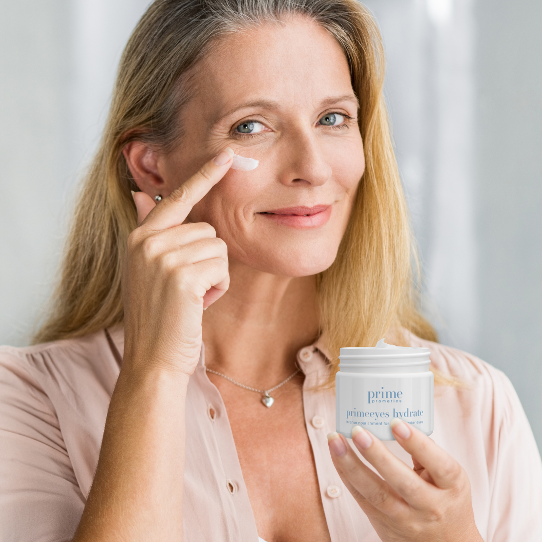 A smiling woman with long blonde hair applies cream under her eye with one finger while holding a jar labeled prime eyes hydrate. She is wearing a light pink blouse and a heart-shaped necklace.