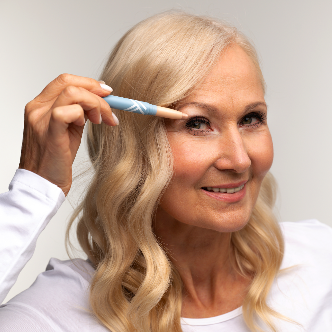 A smiling older woman with long blonde hair applies makeup around her eye using a blue makeup pencil. She is wearing a white top and posing against a neutral background.