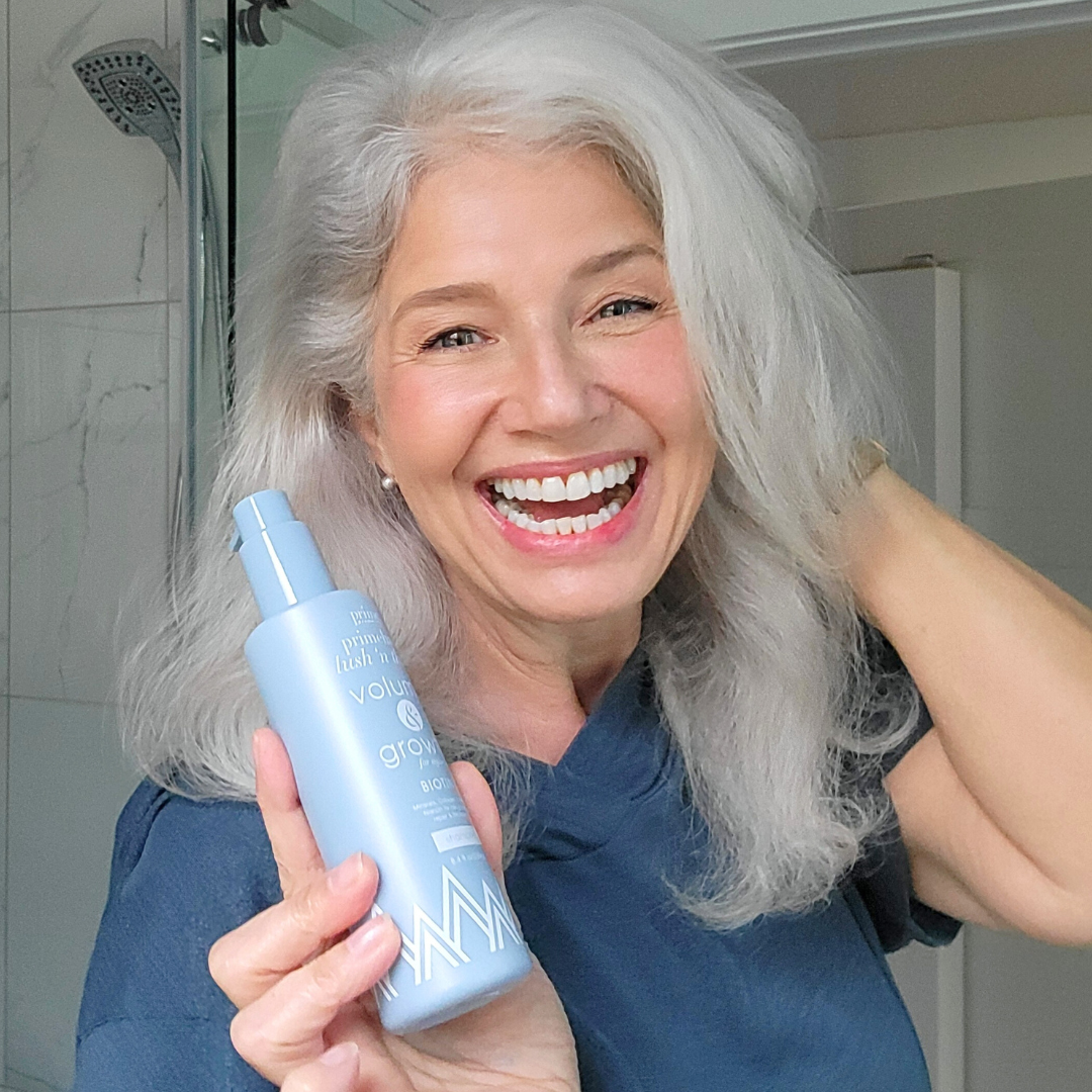 Smiling woman with long gray hair holds up a light blue bottle of hair product in a bathroom, appearing happy and confident as she shows off her healthy hair.
