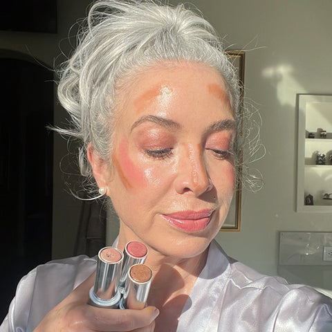 A woman with gray hair holds three makeup sticks against her chin. She has contour, blush, and highlight makeup applied to her face, and is standing in bright natural light.