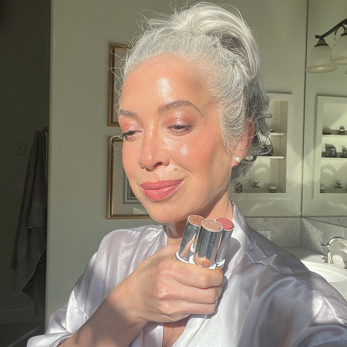 A woman with gray hair in a bun, wearing a light satin robe, holds three lipsticks and smiles in a sunlit bathroom. The sunlight highlights her glowing skin and soft makeup. Two framed pictures and a towel are visible in the background.