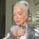 A woman with gray hair in a bun, wearing a light satin robe, holds three lipsticks and smiles in a sunlit bathroom. The sunlight highlights her glowing skin and soft makeup. Two framed pictures and a towel are visible in the background.