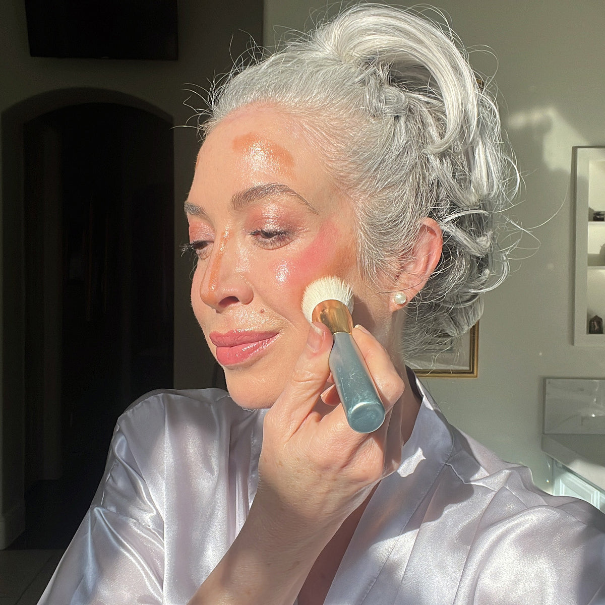 A woman with white hair in an updo, wearing a satin robe, gently applies PrimePrometics™ PrimeWand Contour Kit bronzer to her cheek with a brush in a sunlit room, smiling softly and looking relaxed.