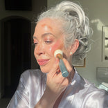 A woman with white hair in an updo, wearing a satin robe, gently applies PrimePrometics™ PrimeWand Contour Kit bronzer to her cheek with a brush in a sunlit room, smiling softly and looking relaxed.