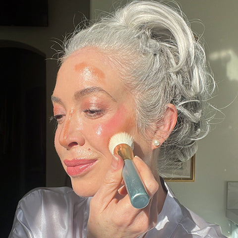 An older woman with gray hair in an updo applies makeup with a brush to her cheek. She has luminous, highlighted skin and is smiling softly, wearing a satin robe, in bright natural light.