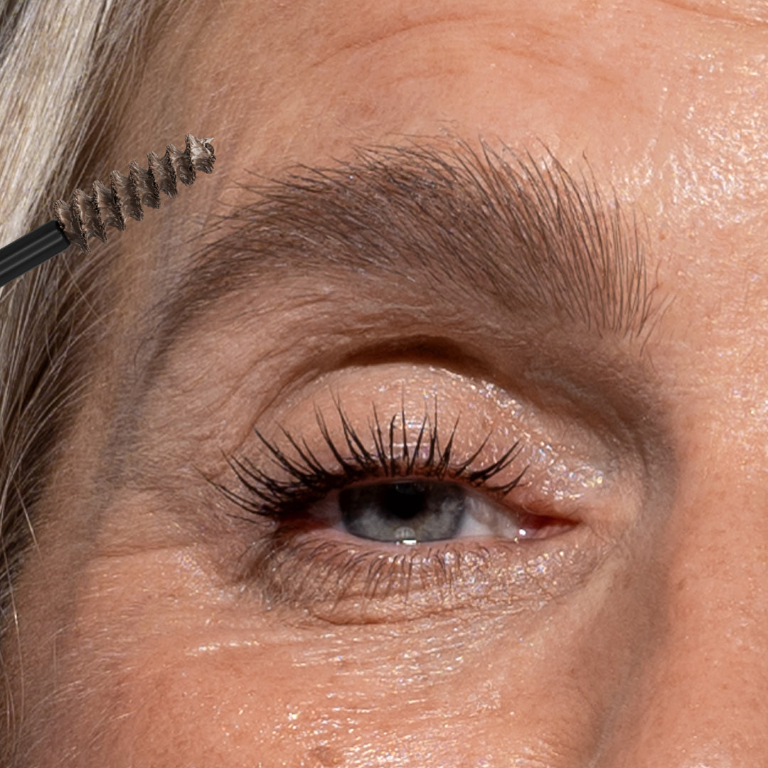 Close-up of an older persons eye and eyebrow as a spoolie brush is used to groom the eyebrow hairs. The skin shows natural texture and fine lines.