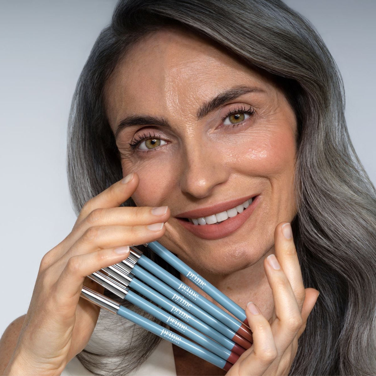 A smiling woman with long gray hair holds several blue makeup pencils by PrimePrometics™ Soft Lip Duo near her face against a plain light background, showcasing the smooth finish of the product.