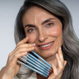 A smiling woman with long gray hair holds several blue makeup pencils by PrimePrometics™ Soft Lip Duo near her face against a plain light background, showcasing the smooth finish of the product.