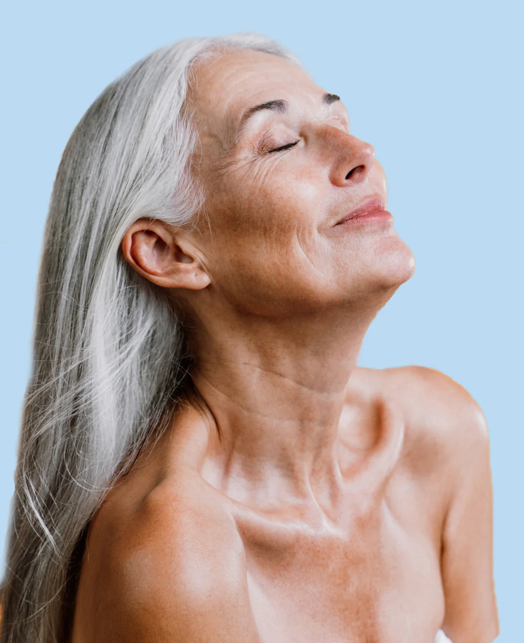 Older woman with long gray hair and bare shoulders, eyes closed and head slightly tilted back, smiling contentedly against a light blue background.