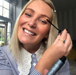 A smiling woman with blonde hair applies mascara to her eyelashes while looking into the camera. She is wearing a striped shirt with a white lace collar and gold jewelry, and is indoors near a window.