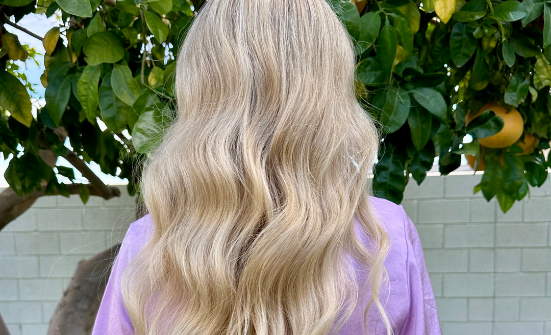A person with long, wavy blonde hair wearing a lavender shirt stands outdoors in front of a white brick wall and green leafy tree. The image is taken from behind.