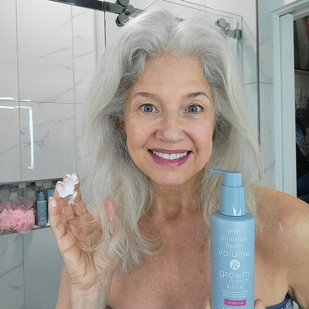 Smiling older woman with long gray hair holds up a dollop of conditioner in one hand and a blue bottle labeled “Prime Luster Volume Growth Conditioner” in the other, standing in a bathroom with tiled walls.