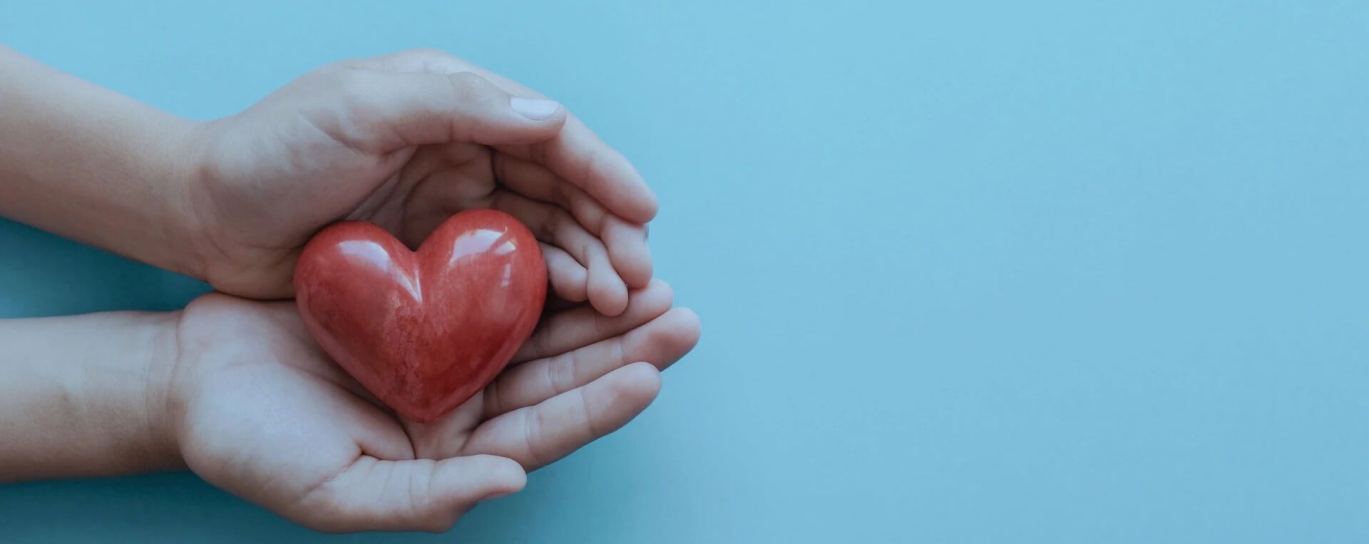 Two hands gently hold a red heart-shaped object against a light blue background, symbolizing care, love, or support.
