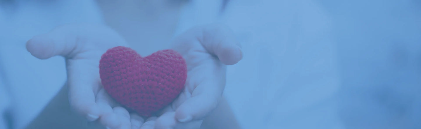 A person holds a small red knitted heart gently in their open hands, symbolizing care and kindness. The image has a soft blue overlay.
