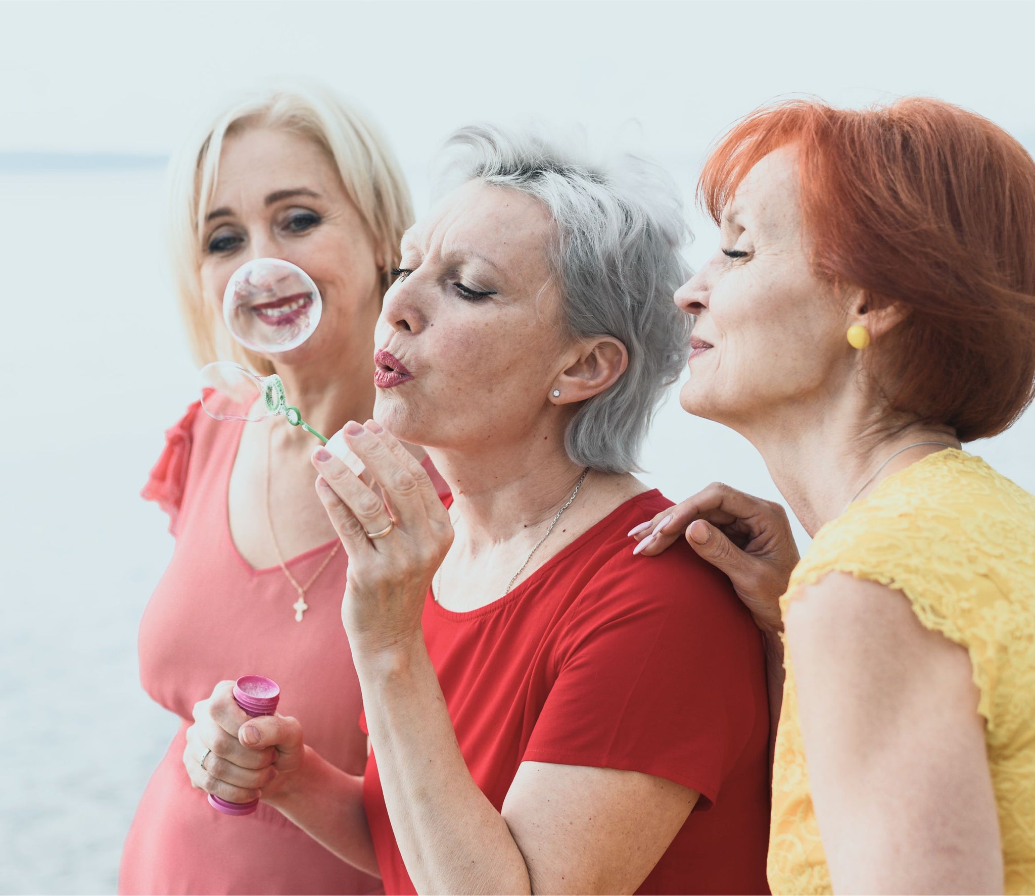 Three older women stand close together outdoors, smiling and laughing as one of them blows bubbles. They appear joyful and relaxed, enjoying each others company. The background is bright and softly blurred.