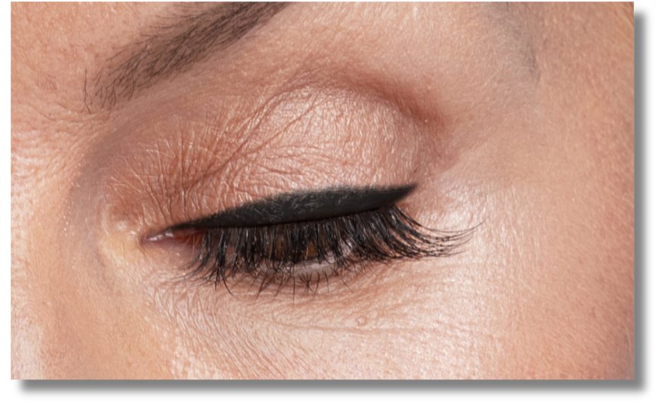 Close-up of a persons closed eye with well-defined black eyeliner and long, dark eyelashes. The skin shows natural texture and a neatly shaped eyebrow is visible above the eye.