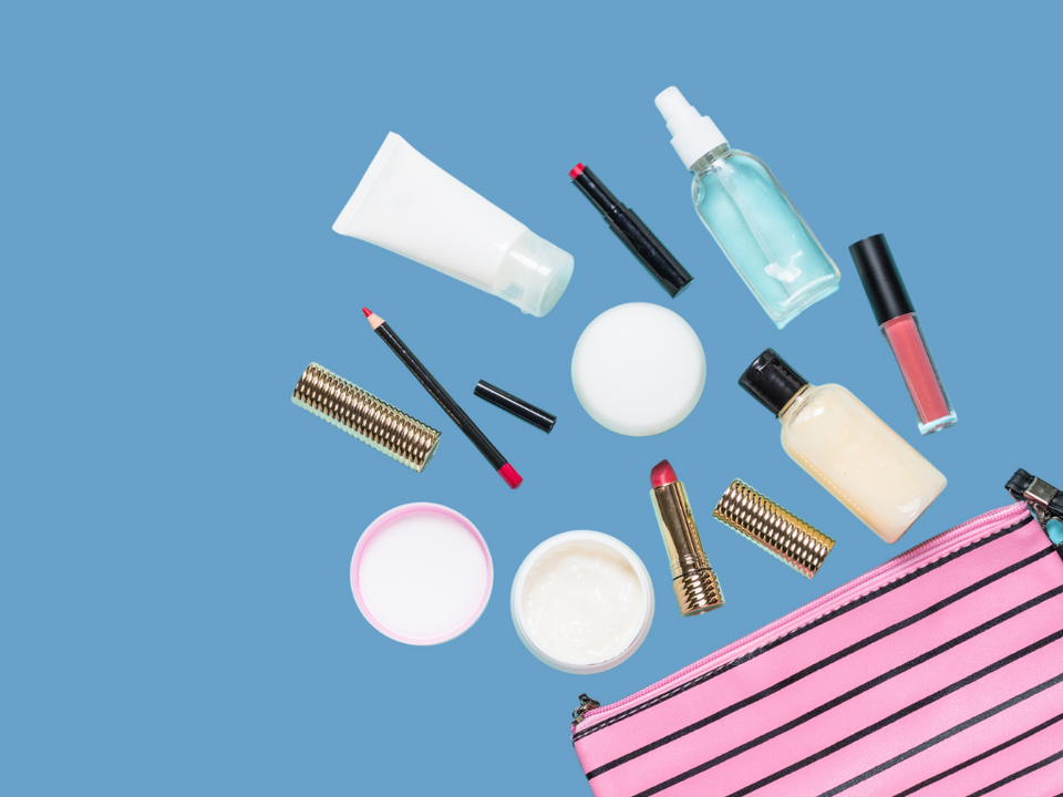 A flat lay of various cosmetics, including lotions, lipsticks, lip gloss, lip liner, and a spray bottle, arranged on a blue background next to a pink striped makeup bag.