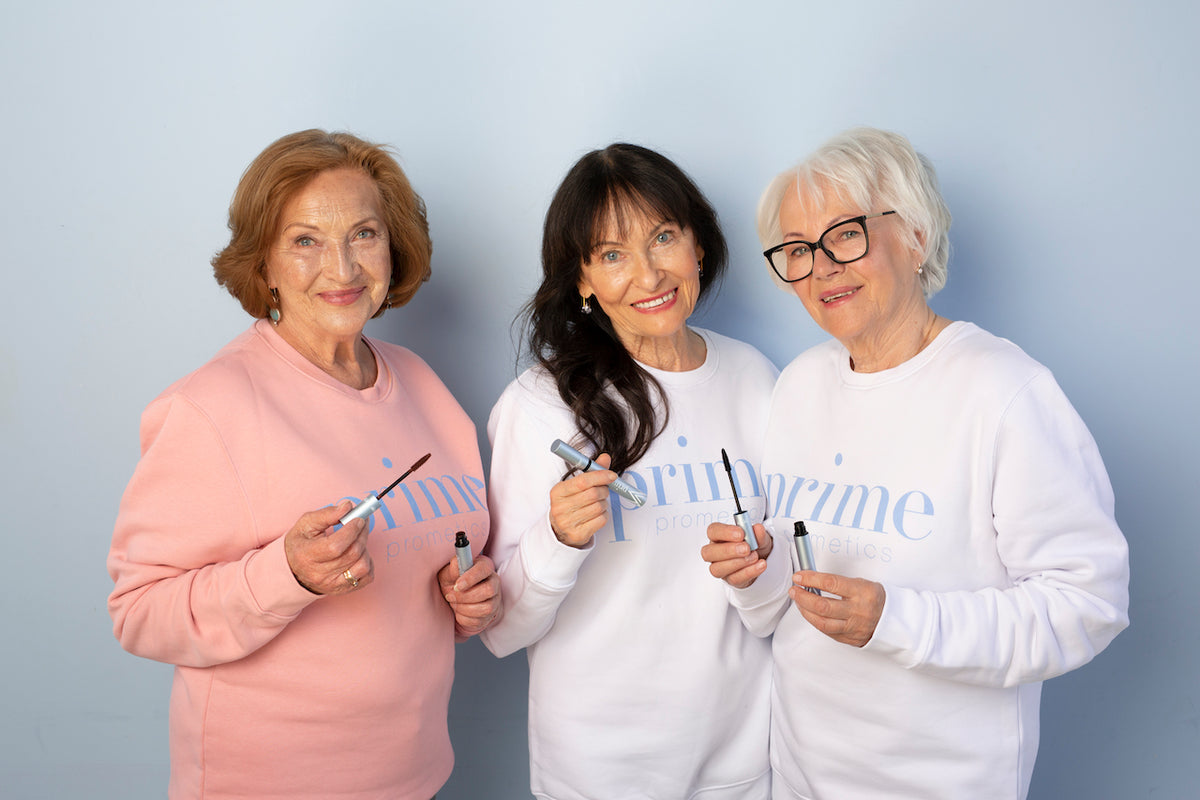 Three older women smiling and holding makeup products, standing together against a light blue background. They are wearing pastel sweatshirts with prime written on them.
