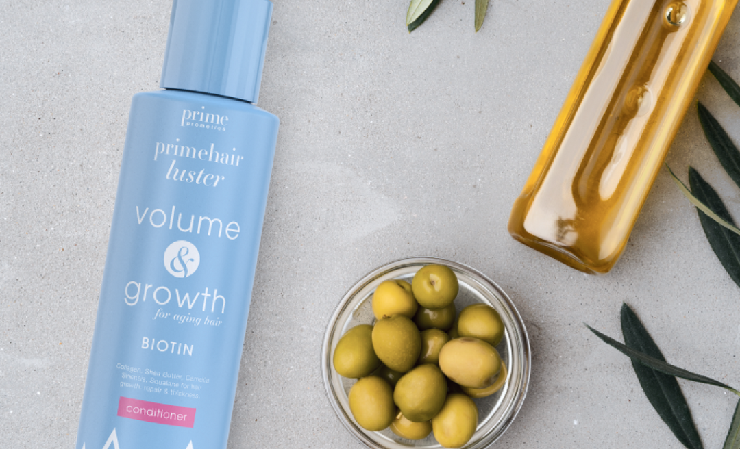 A blue bottle of Prime Hair Luster conditioner, a glass bowl of green olives, a yellow bottle, and olive leaves arranged on a light grey surface.